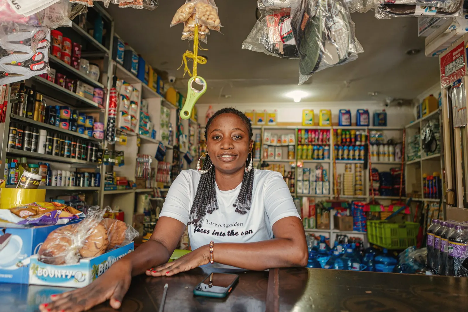 A shop owner leans on the counter of her provisions store, shelves stocked behind her